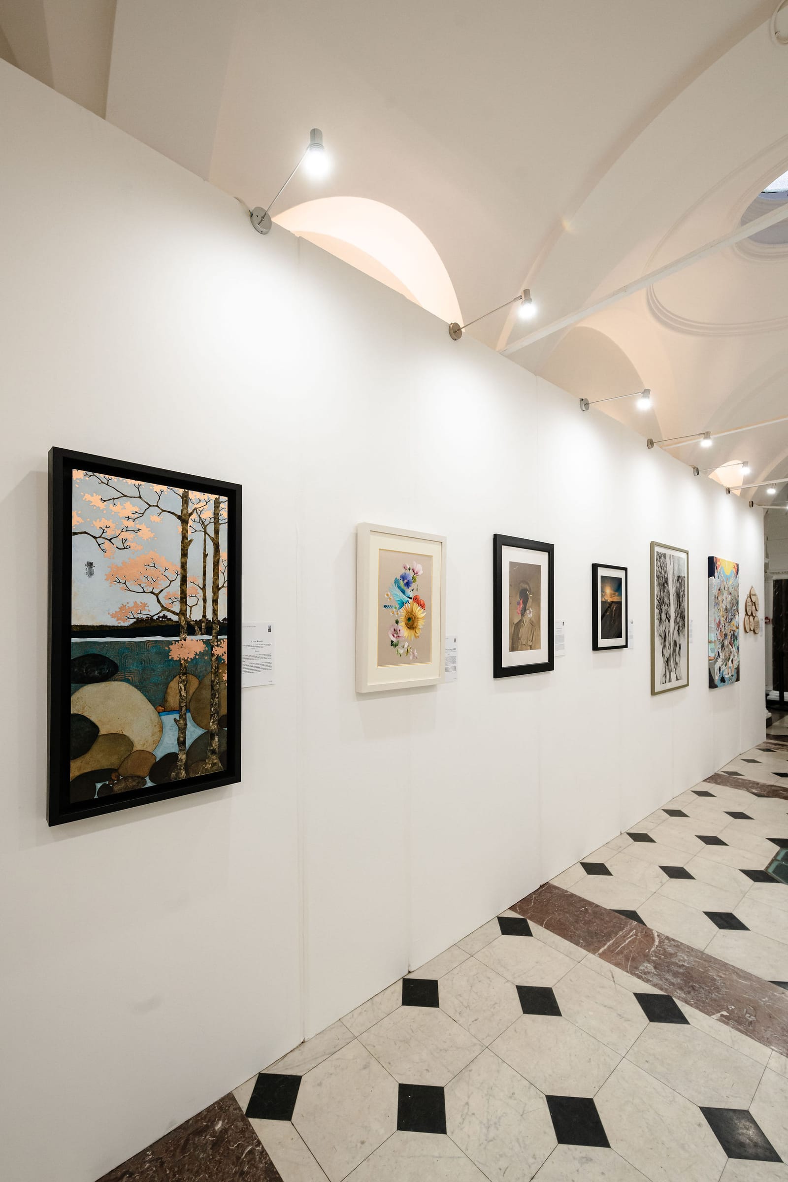 A corridor of the exhibition: framed and unframed works hung along a white wall under gallery lighting, the chequered floor running away beneath them.
