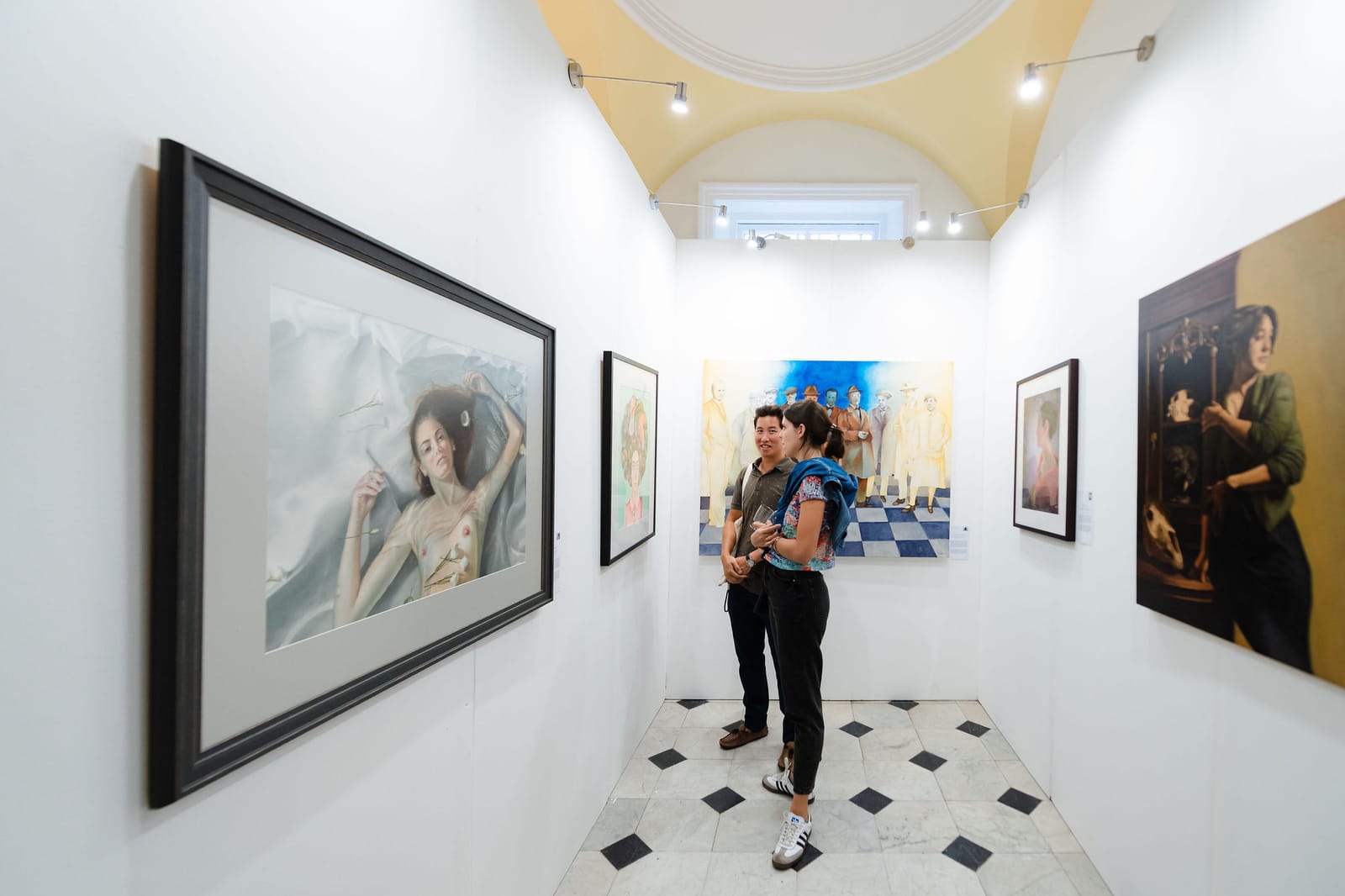 Two visitors standing in a white-walled room of the Biennale, framed works on every wall and a chequerboard floor between them.