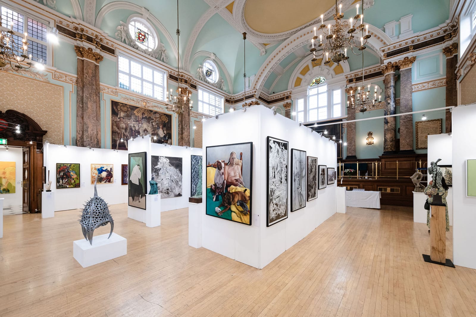 The exhibition hall at Chelsea Old Town Hall, its chandeliers hanging over temporary white walls hung with paintings and a sculpture on a plinth.