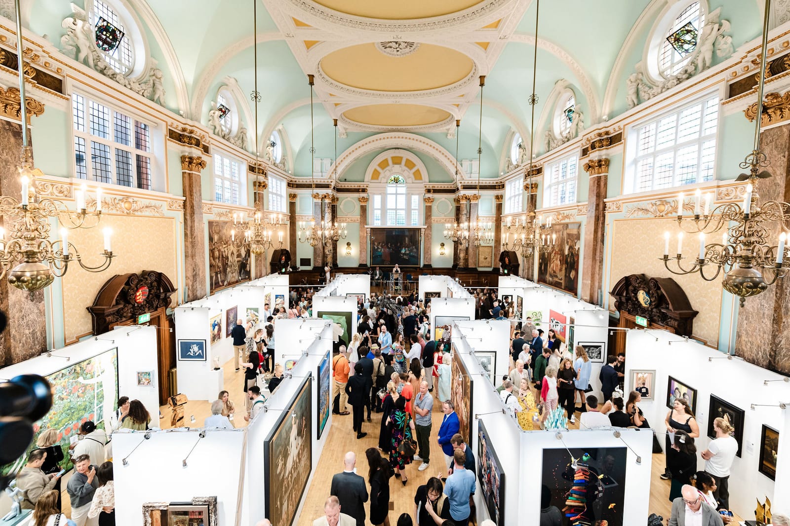 The exhibition seen from the balcony: temporary white walls in a grid across the floor of the hall, visitors moving between them.