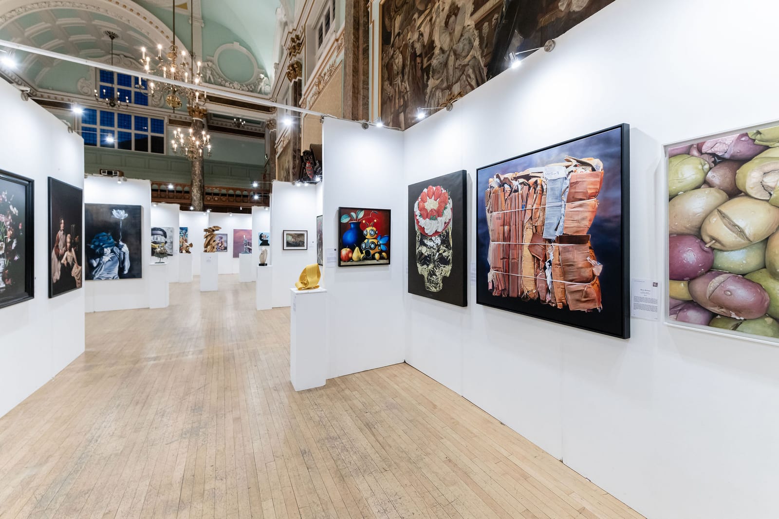 A wide view of the exhibition: temporary white walls hung with paintings of many styles, set within the pale teal and gilded hall of Chelsea Old Town Hall.